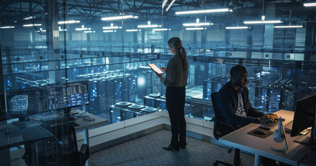 man on computer and woman standing with tablet overlooking a server room - data hosting services