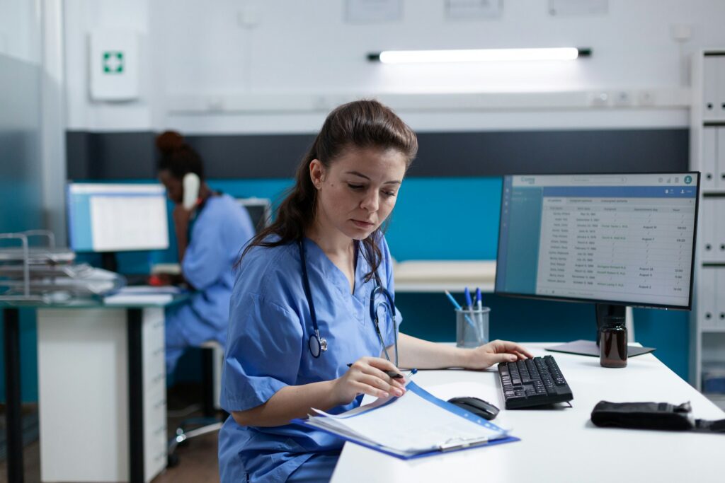 A healthcare worker writing on a clipboard and using a computer.